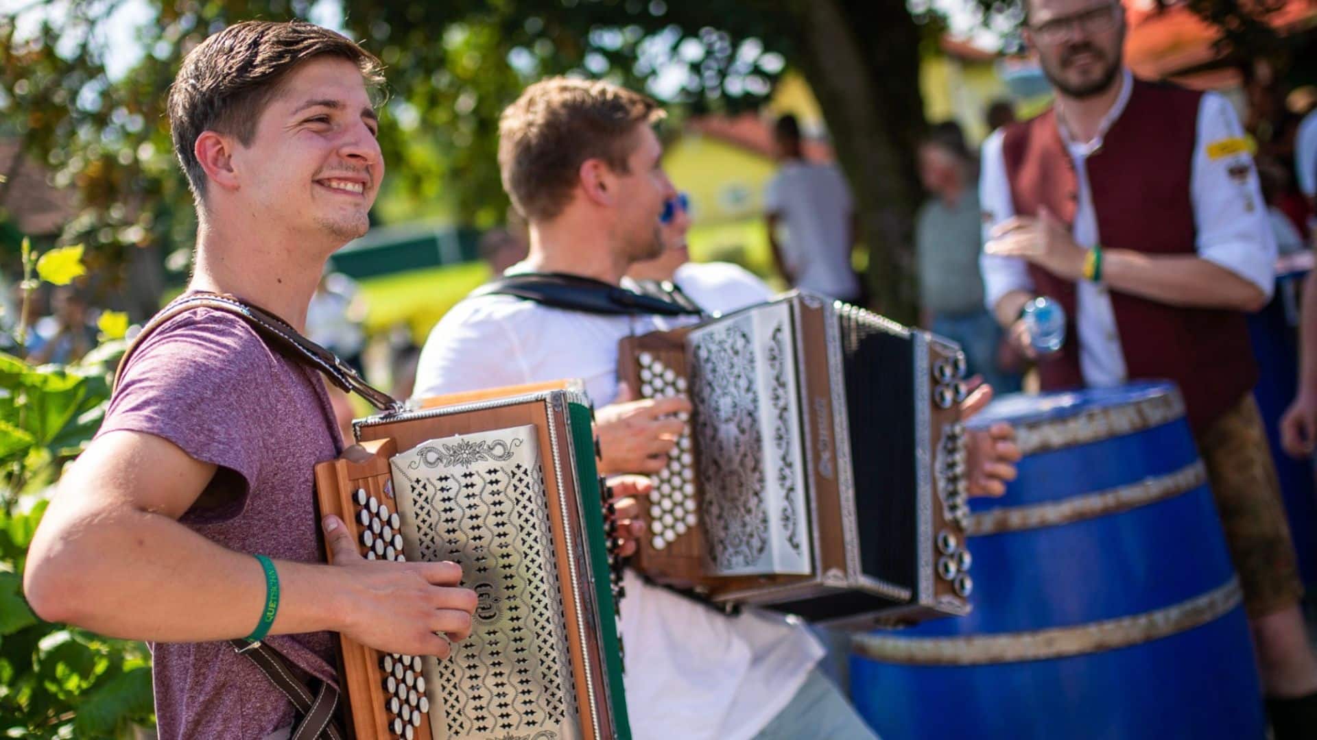 Ein junger Mann im lila T-Shirt spielt draußen lächelnd Harmonika. Hinter ihm spielt ein weiterer Musiker und rechts klatscht ein Mann in Tracht. Die fröhliche und gesellige Szene, die an einen gemütlichen Frühschoppen erinnert, verdeutlicht die Gemeinschaft, die beim Harmonika spielen mit anderen entsteht.
